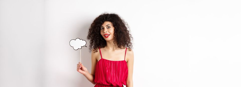 Beautiful woman with curly hair, wearing red dress, holding comment cloud, say something, standing on white background.