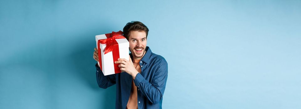 Valentines day. Happy young man got present on special holiday, trying guess what inside gift box and smiling, standing over blue background.