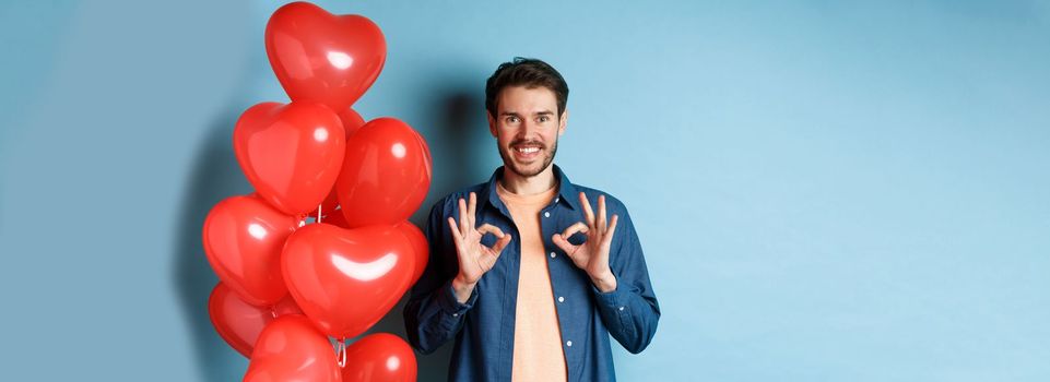 Happy valentines day. Cheerful boyfriend showing okay gestures and praise something good, standing with red hearts balloons for lover, blue background.