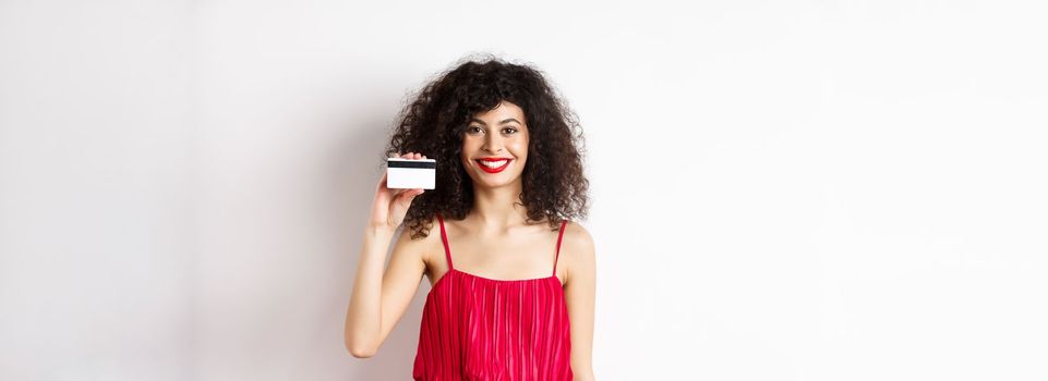 Beautiful woman in red trendy dress and makeup, showing plastic credit card and smiling, recommending offer, standing on white background.