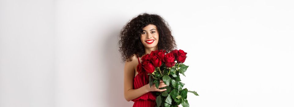 Gorgeous caucasian woman in red evening dress receiving bouquet of flowers. Girl with red roses smiling at camera, having romantic date on Valentines lover day, white background.