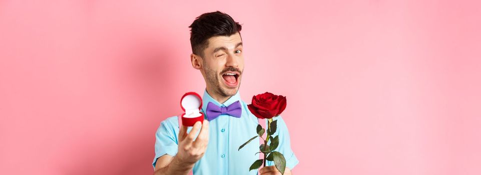 Valentines day. Funny guy making proposal, winking and saying marry me, showing engagement ring with red rose, standing over pink background.