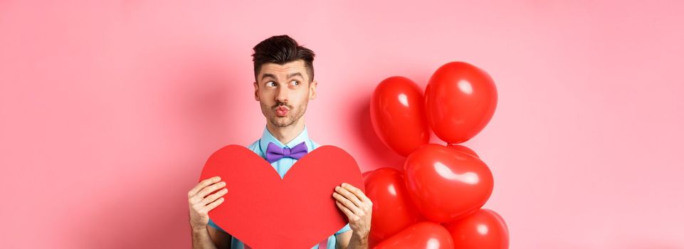 Valentines day concept. Charming young man in bowtie waiting for soulmate with big red heart cutout, looking left and dream of love, pink background.