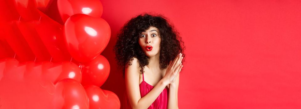 Excited beautiful woman with curly hair, standing near heart balloons and rubbing palms together, expect good deal or relish, red background.