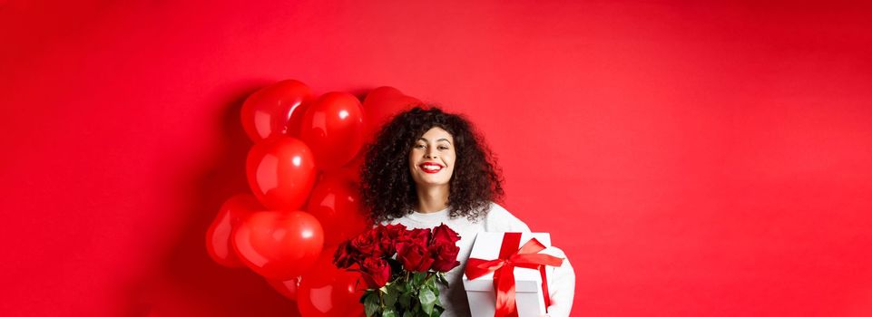 Smiling happy woman holding box with gift and red roses from boyfriend, celebrating Valentines day, standing near romantic hearts balloons, standing over studio background.