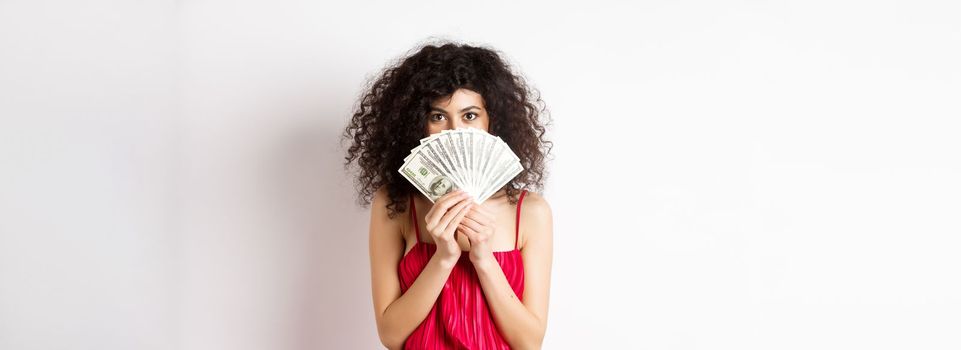 Gorgeous woman with curly hair, hiding face behind dollars and smiling, standing over white background. Copy space