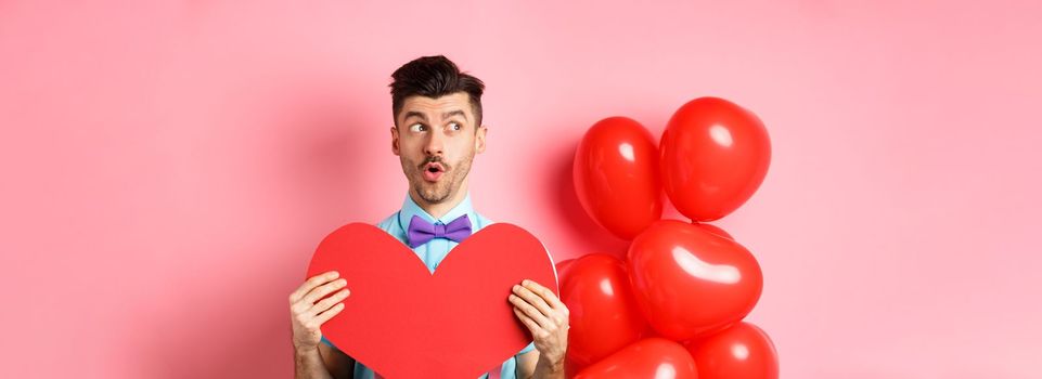 Valentines day concept. Cute young man looking left amused, showing red heart cutout and standing near balloons, pink background.