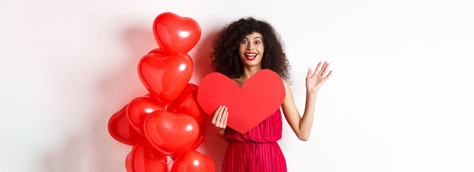 Valentines day and love concept. Cheerful young woman in elegant red dress, standing near romantic balloons and holding big red heart cutout, waving hand to say hi, waiting for date.