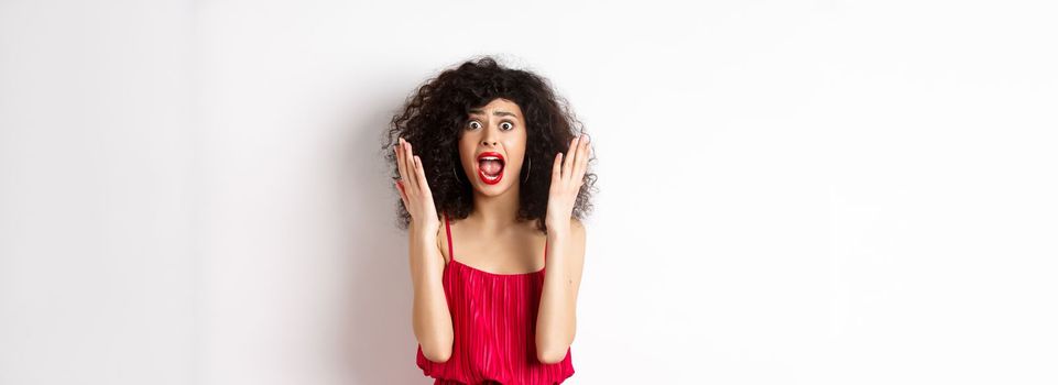 Woman scream in panic, wearing red dress and shouting at camera with anxious face, standing over white background. Copy space