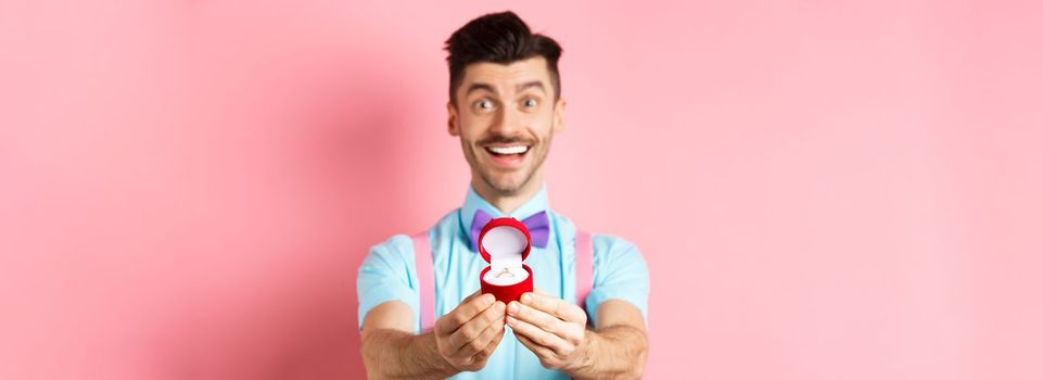 Valentines day. Romantic young man in bow-tie stretch hands with engagement ring and smiling, asking to marry him, making proposal, standing over pink background.