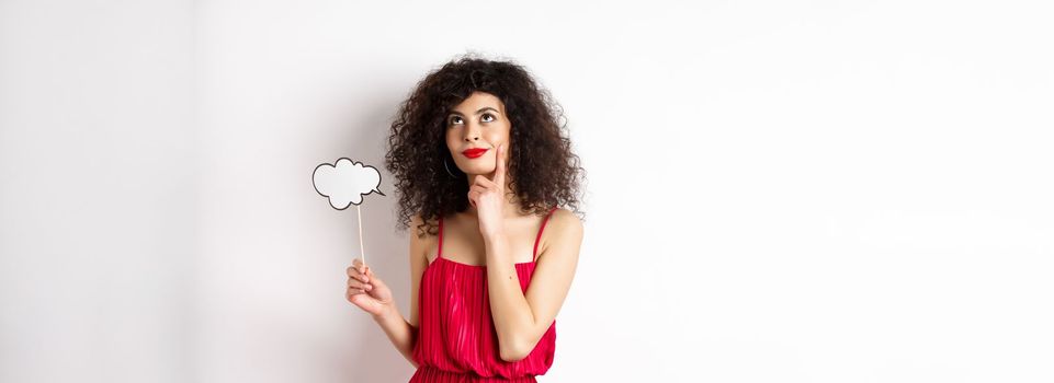 Thoughtful girl in red dress, holding small cloud and looking up with pleased smile, imaging things, standing over white background.