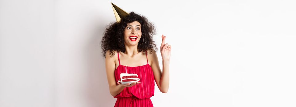 Excited birthday girl making wish, cross fingers good luck and looking aside at logo, celebrating bday, holding piece of cake, white background.