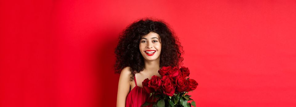 Happy fashionable female model with bouquet of red roses, smiling and looking cheerful at camera, studio background.