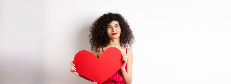 Young romantic woman dreaming about love on Valentines day, looking for soulmate, holding big red heart cutout and gazing pensive left, white background.