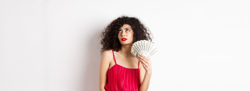 Elegant beautiful woman in red dress, waving with dollars and looking fabulous, standing on white background. Copy space