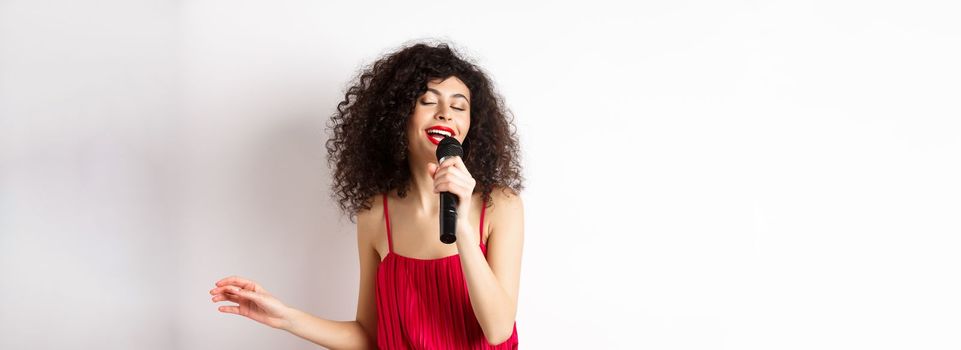 Happy elegant woman in red dress performing with microphone, singing karaoke and smiling, standing over white background.