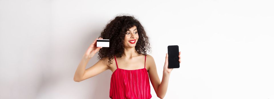 Online shopping concept. Elegant curly-haired woman in red dress showing plastic credit card and empty mobile phone screen, standing over white background.