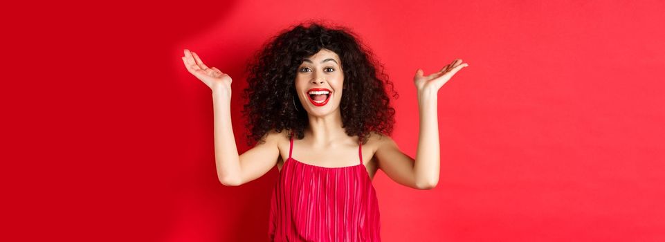 Excited beautiful woman raising hands up and scream with happiness and joy, applause with rejoice, standing on red background.