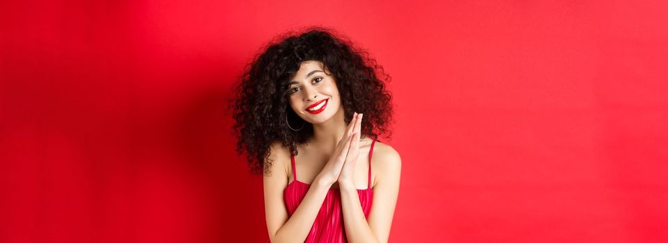 Tender woman with curly hair, wearing red dress, looking at something lovely, thanking you, standing over studio background. Copy space