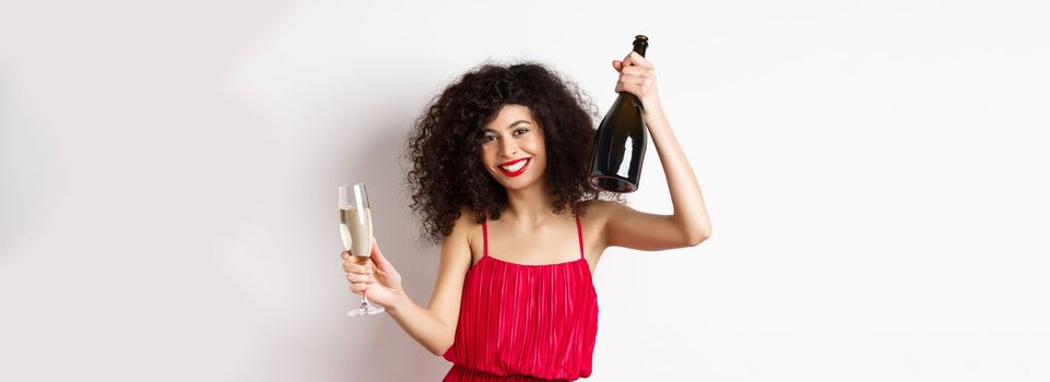 Happy woman partying on valentines day holiday, dancing with glass and bottle of champagne, wearing red dress, smiling on white background.