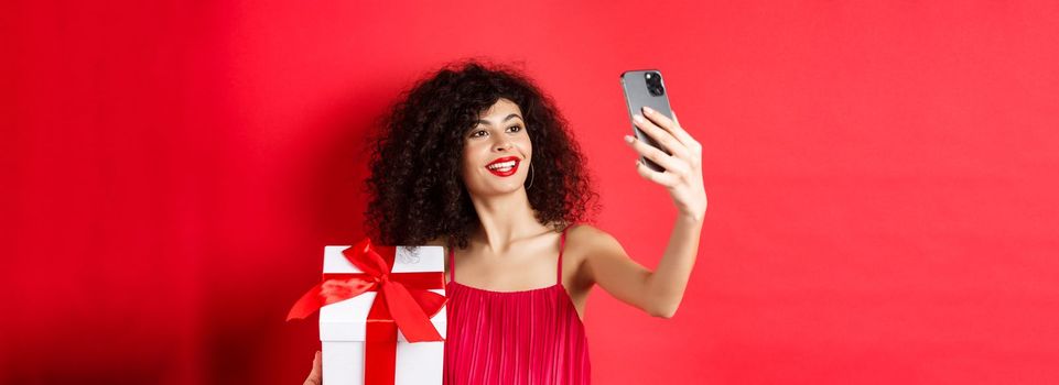 Beautiful girlfriend with curly hair, wearing evening dress, taking selfie with gift from lover, photographing on smartphone and smiling, standing over red background.