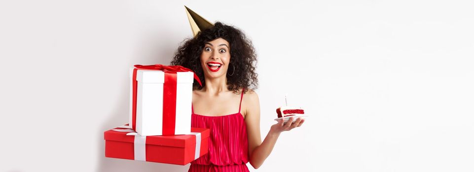 Cheerful girl in party hat and red dress, celebrating birthday, holding surprise gift and b-day cake, enjoying holiday, standing over white background.