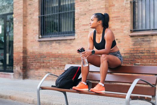 latina female athlete with a bottle of water resting while sitting on a bench during a break in her workout, urban sport and healthy lifestyle concept, copy space for text