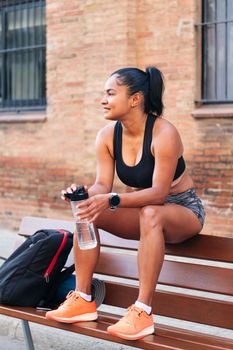 latina female athlete with a bottle of water resting while sitting on a bench during a break in her workout, urban sport and healthy lifestyle concept, copy space for text