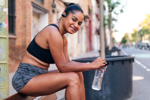 latina sportswoman holding a water bottle smiles while sitting looking at camera during a break in her workout, urban sport and healthy lifestyle concept, copy space for text