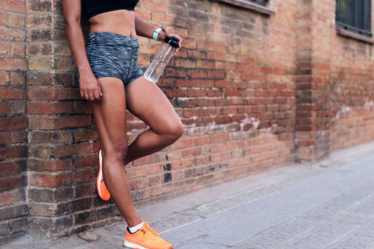 body of an unrecognizable sportswoman holding a bottle of water while resting against a brick wall during a break in her training, urban sport and healthy lifestyle concept, copy space for text