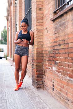 smiling sportswoman using her mobile phone resting against a brick wall during a break in her workout, urban sport and healthy lifestyle concept, copy space for text