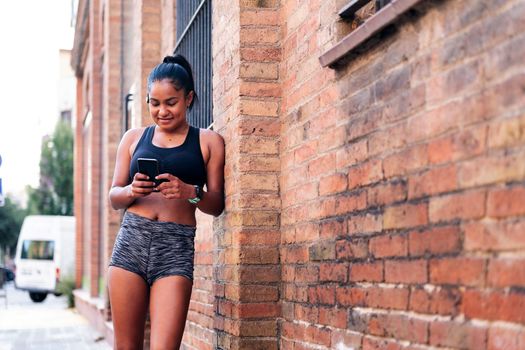 latin sportswoman using her mobile phone resting against a brick wall during a break in her workout, urban sport and healthy lifestyle concept, copy space for text