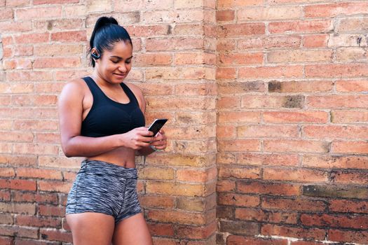 latin sportswoman using her mobile phone resting against a brick wall during a break in her workout, urban sport and healthy lifestyle concept, copy space for text