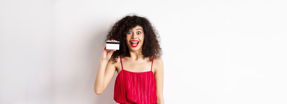 Excited smiling woman in red dress, showing plastic credit card and looking happy, standing over white background. Copy space