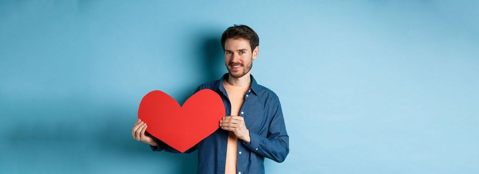 Happy man showing valentines heart and smiling, make romantic gift on lovers day, standing over blue background.