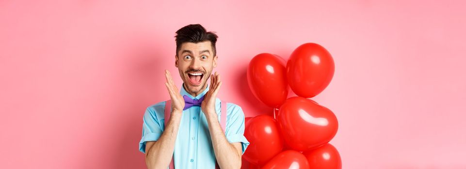 Love and romance concept. Happy man screaming from fantastic news, shouting wow and smiling amused, checking out special offer on Valentines, standing near red hearts balloons.