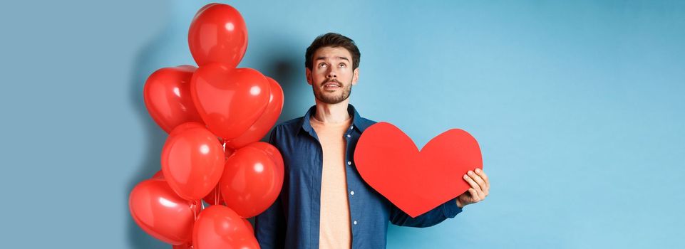 Valentines day and love concept. Man looking up with dreamy face, holding romantic gift balloons and red heart cutout, standing over blue background.