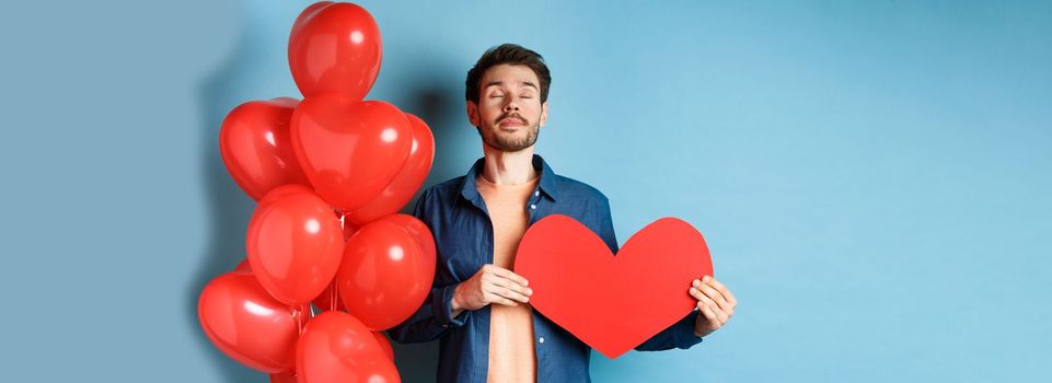 Valentines day concept. Man dreaming of true love, holding red heart cutout and standing near romantic balloons, blue background.