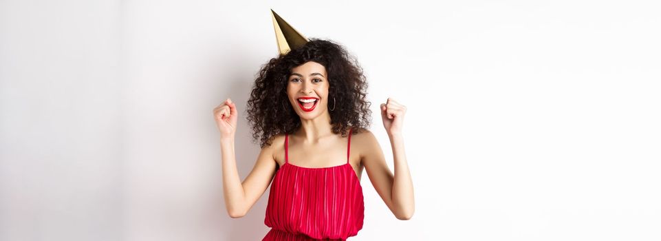 Birthday girl in party hat having fun, dancing in red dress and chanting, standing against white background.