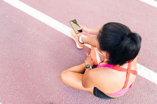 top view of an unrecognizable female athlete using her mobile phone sitting on the athletics track after her workout, concept of sport and healthy lifestyle, copy space for text