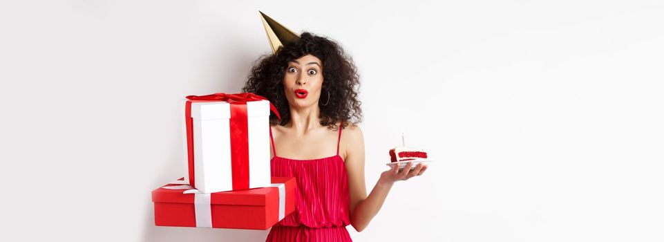 Funny lady in red dress and party hat, celebrating birthday, holding b-day presents and cake with candle, looking amused at camera, standing over white background.