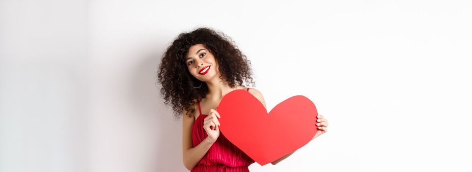 Romantic woman in dress showing big red heart, falling in love, smiling happy at camera, white background.