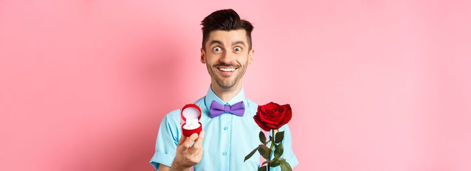 Valentines day. Smiling handsome man asking to marry him, showing engagement ring and red rose, standing romantic on pink background.