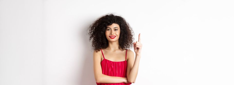 Beautiful smiling lady in red dress showing number one, raising finger and looking pleased, standing over white background.