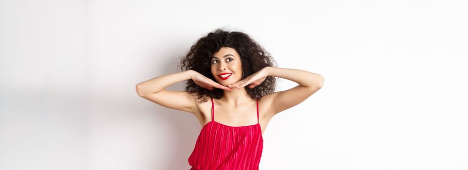 Beautiful lady with curly hair, wearing red dress, showing hear face with makeup and smiling, feeling carefree on white background.