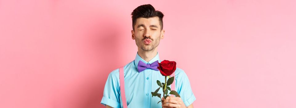 Cute and funny man waiting for kiss from lover on Valentines day, holding beautiful red rose for girlfriend, standing over pink background.