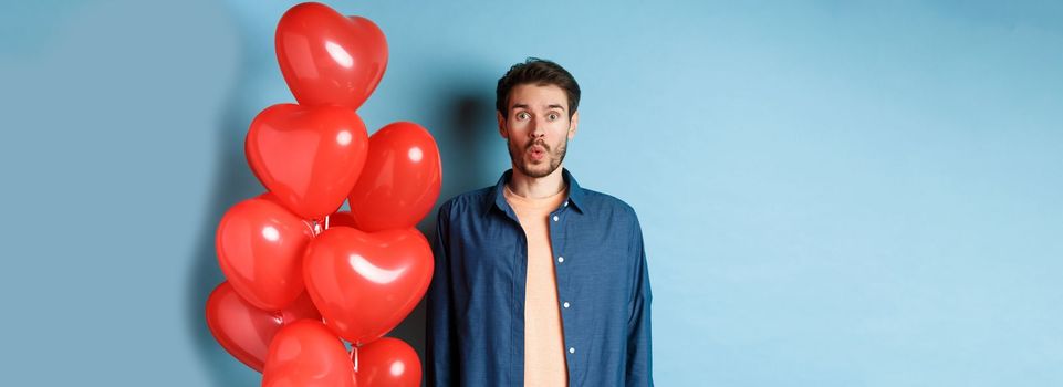 Happy valentines day. Man looking surprised and interested at camera, saying wow, standing near red hearts balloons, blue background.