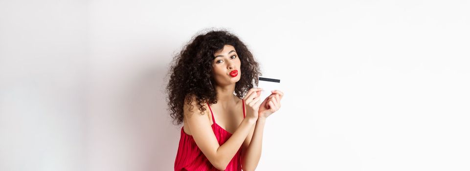 Shopping. Beautiful lady with curly hair, pucker lips, kissing plastic credit card, standing in red dress against white background.