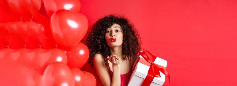Romance and Valentines day concept. Pretty curly-haired girl in red dress sending her love, blowing air kiss at camera, holding gift from lover, standing near hearts on red background.