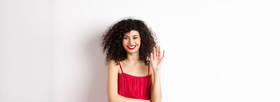 Cheerful elegant woman saying hello, waving hand and smiling at camera, greeting someone, standing in red dress on white background.